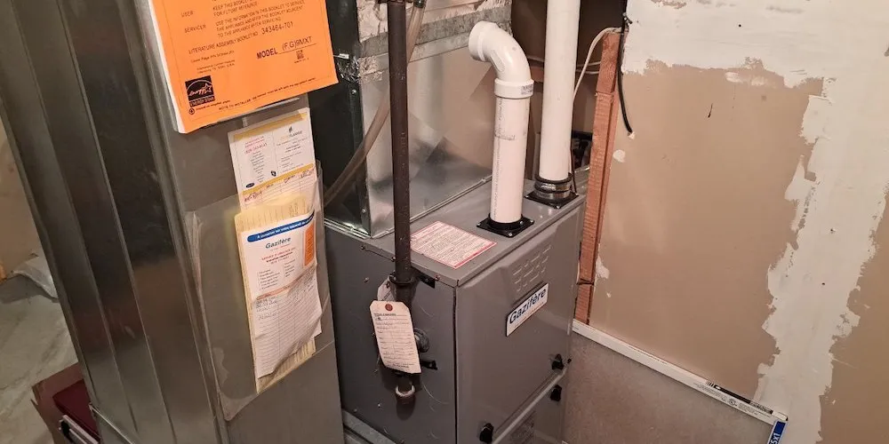 Une vue d'ensemble de l'installation de la fournaise dans une salle mécanique. L'unité est reliée à des conduits métalliques et à des tuyaux d'évacuation en PVC blanc. Des manuels d'entretien jaunes et oranges sont fixés sur le côté.