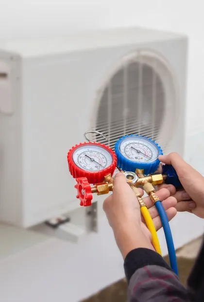 Les mains d'un technicien tenant un jeu de manomètres avec des cadrans rouges et bleus et des tuyaux multicolores (jaune et bleu). Un climatiseur mural blanc est monté sur un mur blanc en arrière-plan.