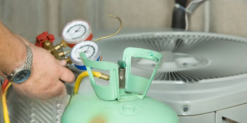Gros plan des mains d'un technicien CVC tenant un collecteur de manomètres (cadrans rouge et bleu) relié à un réservoir de réfrigérant vert clair et à un climatiseur extérieur gris.