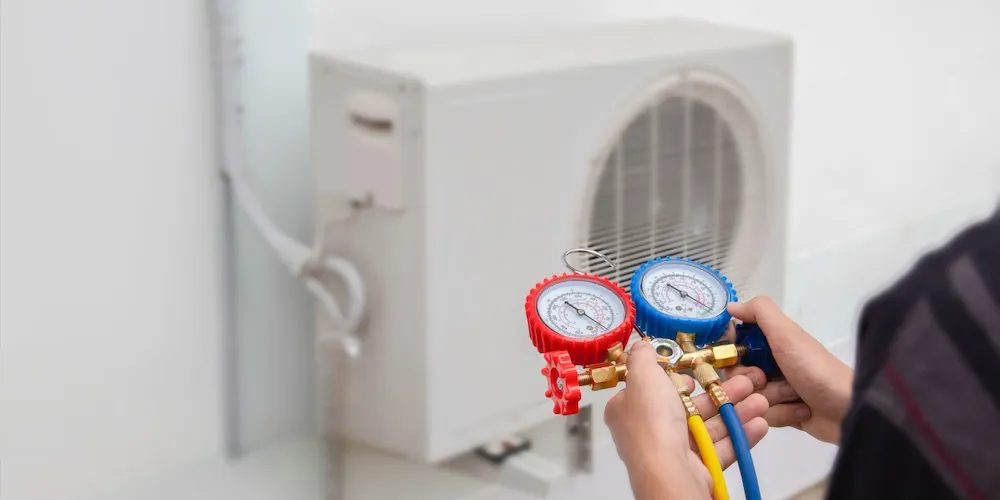 Un technicien tient un jeu de manomètres au premier plan, avec des tuyaux jaunes et bleus connectés, tandis qu'une unité de condensation extérieure de type mini-split blanche est visible à l'arrière-plan.