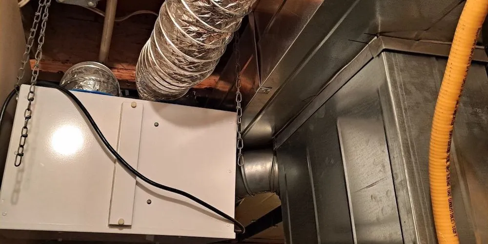 Vue intérieure d'un échangeur d'air rectangulaire blanc suspendu aux solives du plafond par des chaînes métalliques. Il est relié à un gros conduit flexible argenté et à divers conduits de ventilation en métal. Un tuyau de gaz flexible jaune et des câbles électriques sont également visibles.