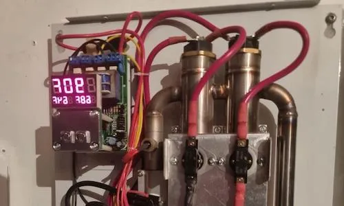 Un gros plan d'une chaudière électrique montrant deux éléments chauffants en acier inoxydable reliés par des fils rouges de gros calibre. À gauche, un affichage numérique à DEL indique la température, et plusieurs relais électriques sont visibles en dessous.