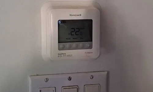 Un thermostat numérique Honeywell ProSeries blanc monté sur un mur au-dessus d'une plaque d'interrupteurs. L'écran affiche une température ambiante de 22,0°C. Un numéro de service local pour « Gazifère » est inscrit en bas à gauche.