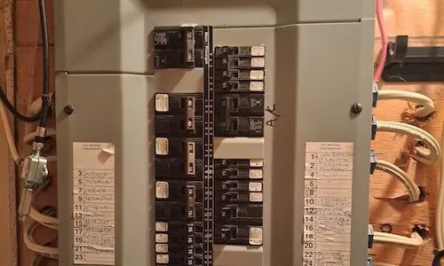 Un panneau de disjoncteurs électriques intérieur sans son couvercle, révélant des rangées de disjoncteurs. Des étiquettes manuscrites des deux côtés identifient les circuits. Un gros disjoncteur bipolaire est marqué « A/C » sur le côté droit.