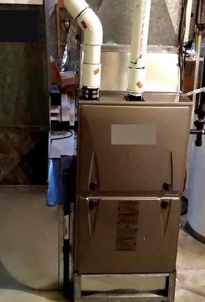 Une vue de face d'une fournaise au gaz à haute efficacité de couleur grise, installée dans un sous-sol. Deux tuyaux d'évacuation en PVC blanc sortent du dessus, et un conduit de retour d'air en métal est fixé sur le côté gauche. L'unité possède deux panneaux d'accès fixés par des vis à oreilles noires.