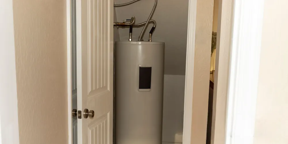 Un chauffe-eau blanc, haut et cylindrique, situé dans un petit placard ou une salle mécanique, avec des conduites d'eau argentées flexibles raccordées au sommet.
