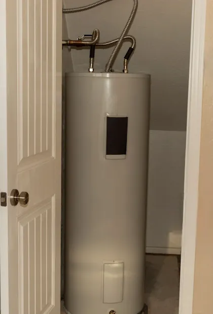 Un chauffe-eau blanc, haut et cylindrique, situé dans un petit placard ou une salle mécanique, avec des conduites d'eau argentées flexibles raccordées au sommet.