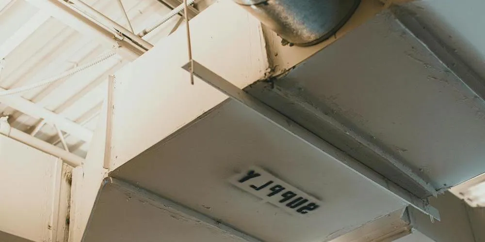 Vue détaillée de grands conduits d'air rectangulaires peints en blanc cassé, montrant les tiges de suspension et un numéro d'identification estampillé sur la surface inférieure.
