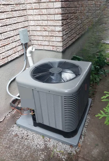 Une vue en plongée d'un condenseur de climatisation central moderne, de couleur grise, installé à l'extérieur sur un socle en plastique gris au-dessus d'une base de gravier. L'unité est placée contre un mur en briques claires ; on aperçoit les conduites de réfrigérant en cuivre, un tuyau d'évacuation en PVC blanc et un boîtier de déconnexion électrique fixé au mur.