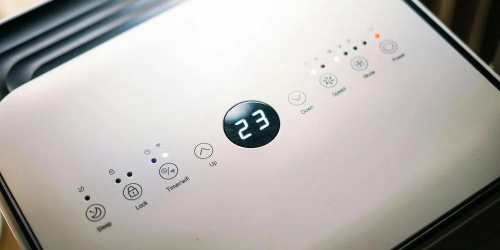 Une vue rapprochée en plongée du panneau de commande tactile d'un climatiseur portable ou d'un déshumidificateur blanc. L'écran numérique au centre affiche le nombre « 23 » en chiffres LED blancs brillants. Autour de l'écran se trouvent des icônes tactiles pour l'alimentation, le mode, la vitesse du ventilateur, la minuterie/Wi-Fi, le verrouillage et le mode nuit.