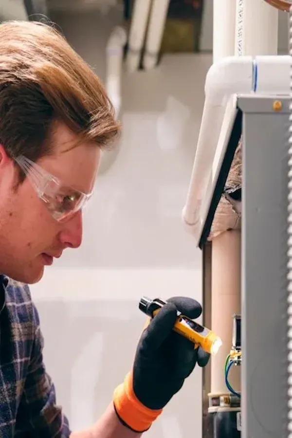 Gros plan de profil d'un technicien portant des lunettes de sécurité et des gants de travail noirs. Il utilise une petite lampe de poche jaune pour inspecter soigneusement le câblage interne et les composants d'un panneau électrique ou CVC gris ouvert.