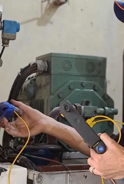 Vue rapprochée des mains d'une personne effectuant la maintenance d'un gros compresseur industriel vert. Une main tient un appareil de test électronique noir avec des fils jaunes, tandis que l'autre main ajuste un connecteur bleu sur un tuyau.